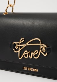 Svart lærveske med et gull "love"-logo, glatt tekstur og en kjede-straps. Pyntet med et merkevaretag nederst.