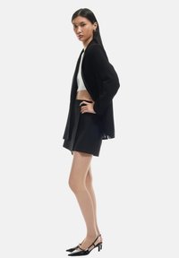Blazer negro sobre una camiseta corta blanca, combinado con una falda negra de cintura alta y zapatos de tacón negro con tira en el talón. Textura suave, ajuste entallado.