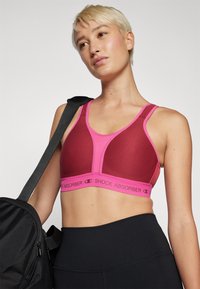 Sujetador deportivo rosa con diseño de malla, tirantes en contraste y una banda elástica de marca. La modelo sostiene un bolso negro junto a unos leggings negros.