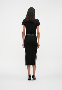 Melnā pieguļošā T-kreklā un ribotā midi svārka ar augstu šķēlumu. Redzamā zīmola apzīmējums jostasvietā. Modelis valkā melnus augstpapēžu apavus.