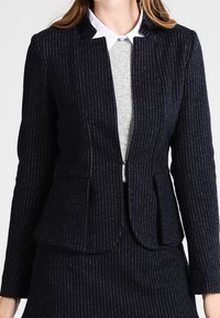 Tummansininen pinstripe-kuvioitu bleiseri, joka on valmistettu teksturoidusta kankaasta ja jossa on istuva leikkaus, ilman kauluksia ja kaksi etutaskua. Päällä harmaa paita.