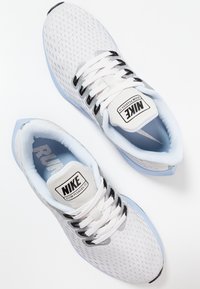 Scarpe da corsa in rete bianche con accenti blu chiaro, cinturini neri e linguetta imbottita. Presentano il marchio "Nike" e dettagli di superficie testurizzati.