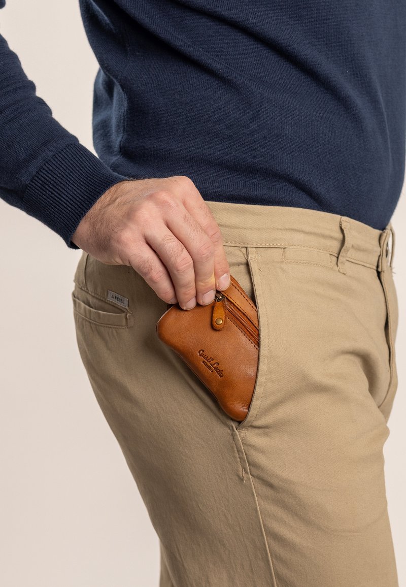 Portefeuille en cuir marron avec fermeture éclair, placé dans une poche de pantalon beige. Le portefeuille présente un logo et un bouton décoratif.