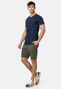 Mørkeblå V-hals t-shirt, olivengrønne shorts og hvide sneakers med gule og sorte detaljer. Shortsene har synlige stikninger.
