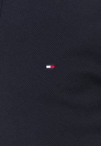 Petit logo rectangulaire avec des blocs de couleur horizontaux en marine, blanc et rouge sur un tissu texturé bleu foncé.