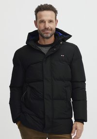 Sort puffjakke med hætte, med en quiltet struktur, sidelommer og blå foring, lukkes med knapper foran.