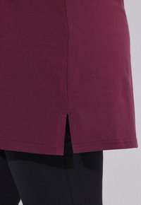 Bordeauxfarbene Tunika mit kurzen Seitenschlitzen, aus weichem Stoff, mit geradem Saum und nahtlosem Design, kombiniert mit schwarzen Leggings.