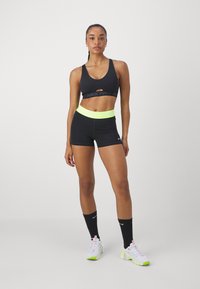 Zwarte sportbeha met uitsnijding en logo, gecombineerd met zwarte shorts met hoge taille en een neongele tailleband en logo. Witte sneakers met neonaccenten.