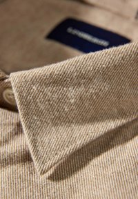 Beige skjorta med krage och ett subtilt randigt mönster, bomullstextur och en liten knapp. Märke synligt nära kragen.