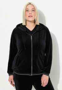 Sudadera con cremallera negra de terciopelo, con capucha ajustable, puños a juego y cremallera plateada. Textura suave con un brillo sutil.