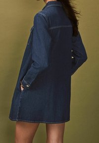 Tmavě modrá džínová košilová šaty s dlouhými rukávy, předními kapsami a uvolněným střihem, sahající po střední stehna. Hladká textura a jednoduchý design.