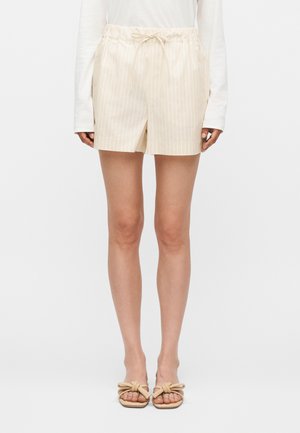 Γυναίκα που φοράει ανοιχτό μπεζ ριγέ σορτς με ελαστική μέση και κορδόνι, σε συνδυασμό με λευκό μακρυμάνικο τοπ και μπεζ σανδάλια με κόμβους.