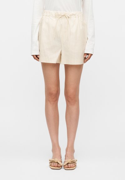Femme portant un short beige clair rayé avec taille élastique et cordon, associé à un haut blanc à manches longues et des sandales beige nouées.