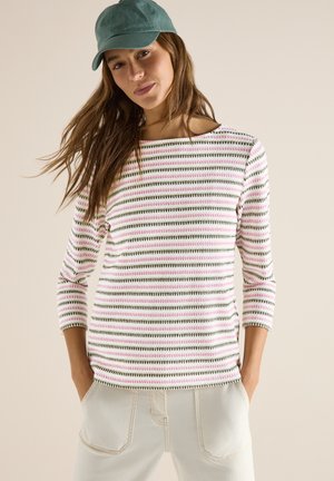 Vrouw met lang bruin haar, een groene pet, een gestreept roze en groen shirt met lange mouwen en lichtbeige broek met handen in de zakken.