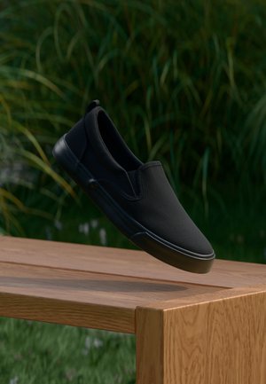 Schwarzer Slip-On-Schuh schwebt über einer Holzbank mit unscharfem, natürlichem grünem Gras im Hintergrund.