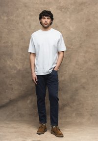 Uomo con capelli scuri e ricci è in piedi davanti a uno sfondo beige testurizzato, indossa una camicia bianca a maniche corte, pantaloni scuri e scarpe marroni.