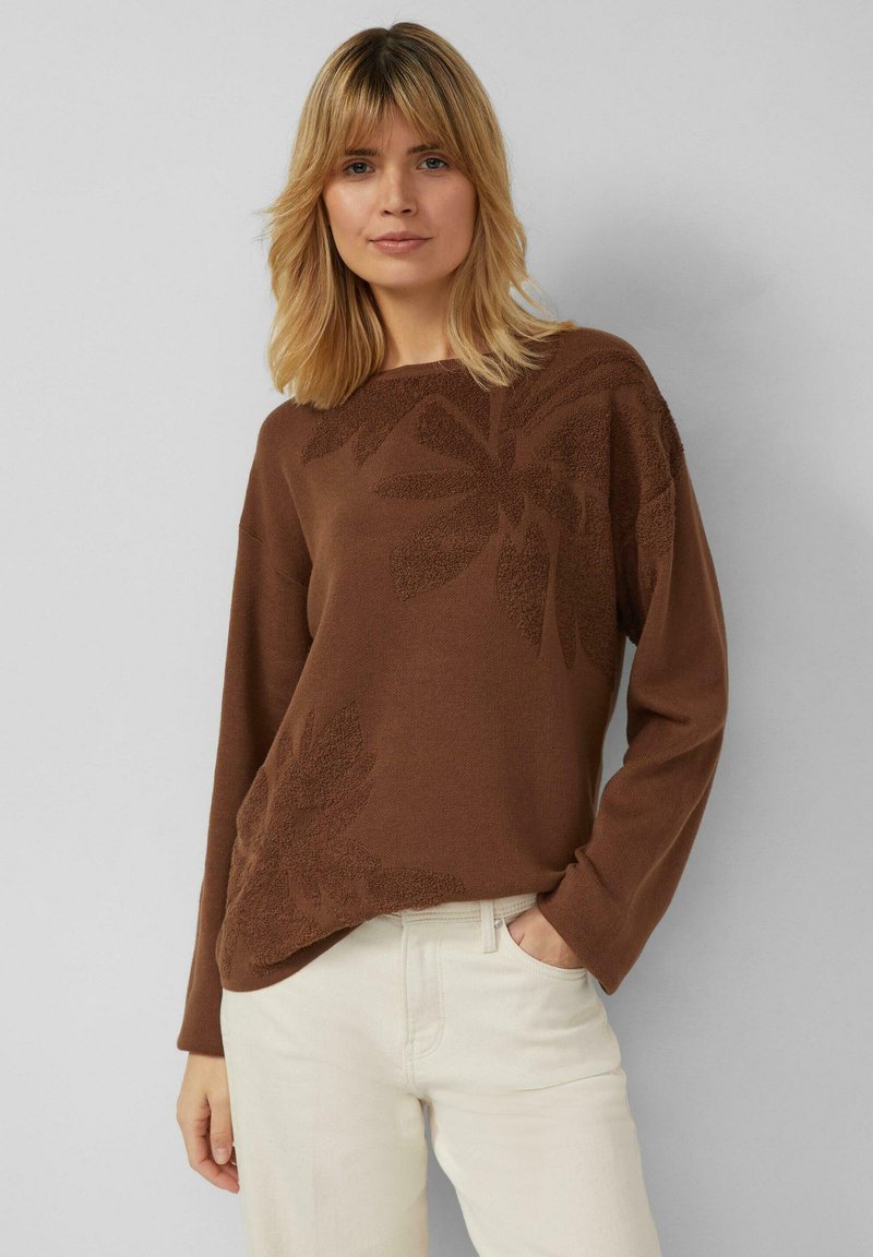 Pull en tricot marron à manches longues, motif floral en nuances de marron plus clair. Porté avec un pantalon couleur crème clair.