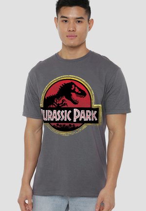 Young man wearing a gray t-shirt with a red, black, and yellow Jurassic Park logo featuring a dinosaur silhouette.