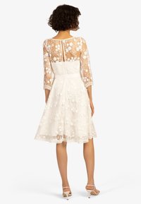 Robe en dentelle blanche avec broderies florales, manches trois-quarts, dos semi-transparent, taille cintrée et jupe évasée, longueur en dessous du genou.