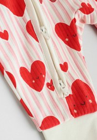 Tessuto rosso con motivo a cuori, strisce rosa e bianche, con chiusura frontale a zip. Texture morbida, con polsini e dettagli a costine.