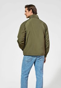 Oliwkowa, lekka kurtka z stójką i elastycznymi mankietami. Połączona z jasnoniebieskimi dżinsami, prezentuje swobodny styl.