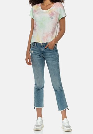 Femme portant un t-shirt tie-dye pastel à manches courtes, un jean bleu clair à ourlet effiloché et des baskets blanches, debout avec une main dans la poche.