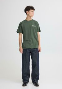 T-shirt en coton vert à manches courtes avec le logo blanc "Soulscape", associé à un jean en denim bleu foncé ample et des chaussures noires.