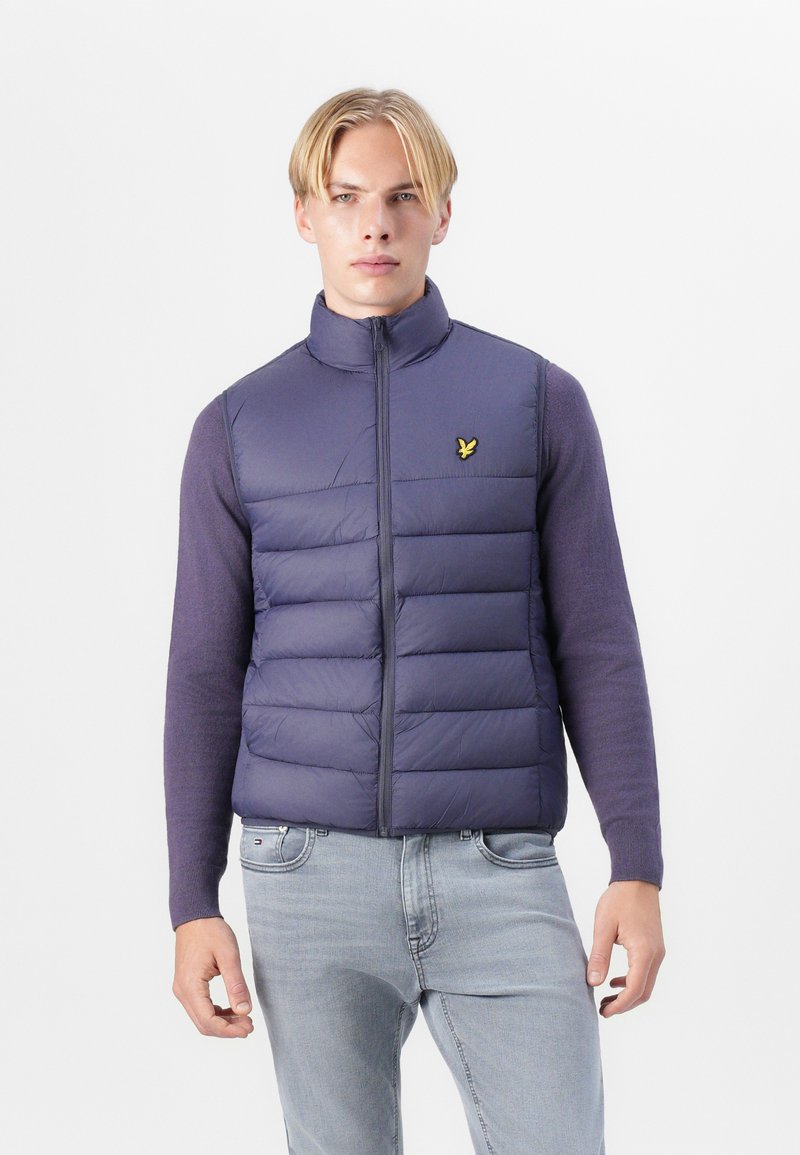 Gilet rembourré violet avec un col haut, présentant des panneaux matelassés et un petit logo jaune sur la poitrine. Porté par-dessus un haut à manches longues.