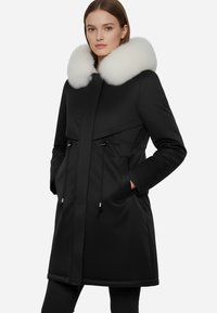 Cappotto nero con colletto in pelliccia sintetica bianca, vita regolabile con cordino e due tasche frontali. Tessuto liscio e opaco con un design elegante e moderno.