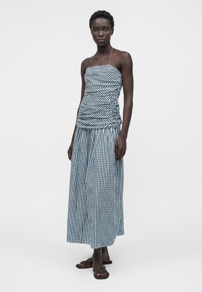 Γυναίκα που φοράει ένα αμάνικο ασπρόμαυρο φόρεμα με καρό μοτίβο, με ρυτιδωτό μπροστινό μέρος και φαρδιά φούστα, σε συνδυασμό με μαύρα σανδάλια τύπου φλίπ-φλοπ, στέκεται όρθια.