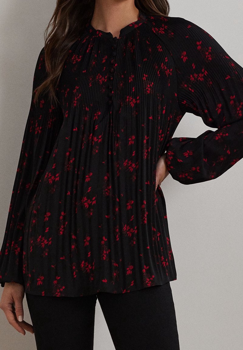 Femme portant un chemisier noir à manches longues avec des motifs floraux rouges et des boutons sur le devant, associé à un pantalon noir.