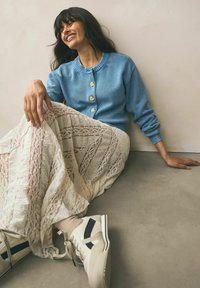 Cardigan tricotat de culoare albastră deschisă, cu trei nasturi aurii, purtat peste o fustă din dantelă albă, cu dungi decorative, asortată cu adidași crem.