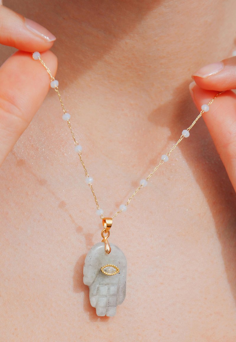 Collier en chaîne dorée avec de petites perles opaques, une pendentif en pierre précieuse grise en forme de main et un détail en forme d'œil en or.