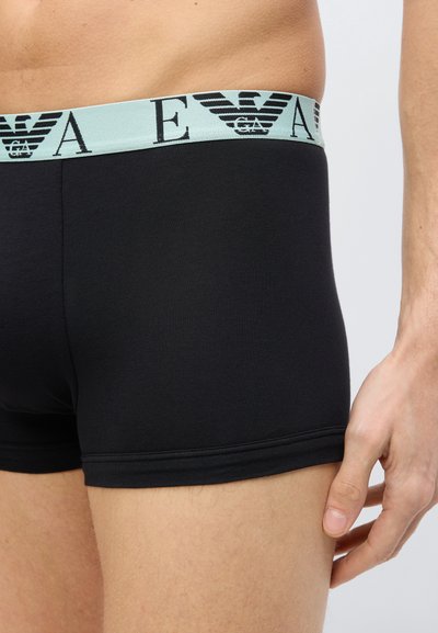 Άνδρας που φοράει μαύρα εσώρουχα boxer με γαλάζια μέση που έχει μαύλο λογότυπο EA και έμβλημα αετού, στέκεται μπροστά σε απλό φόντο.
