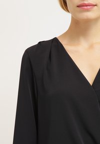 Blouse noire avec un décolleté en V et un détail drapé, fabriquée en tissu lisse ; comporte des manches longues et une coupe fluide.