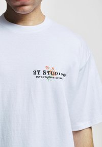 Tricou alb din bumbac, cu un grafic floral mic colorat și textul "2Y STUDIOS" și "INTENTIONAL LIVING" pe partea stângă a pieptului.