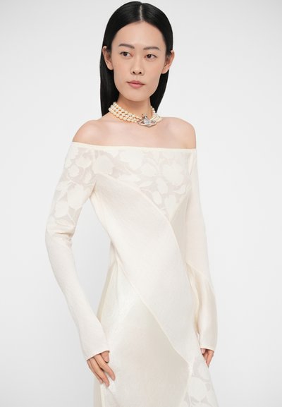 Šaty s odhalenými ramenami a dlhými rukávmi v slonovinovej farbe, zdobené kvetinovou čipkou a textúrovanými panelmi; doplnené perlovým náhrdelníkom s dekoratívnym prvkom.