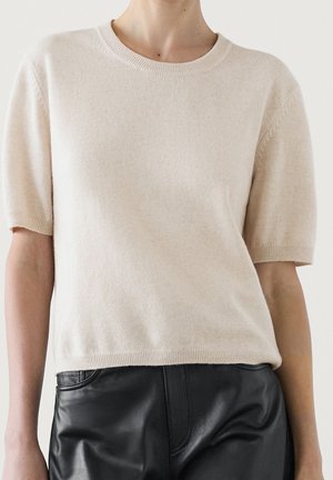 Persoon draagt een beige gebreide trui met korte mouwen, gecombineerd met hoge zwarte leren broeken, tegen een effen achtergrond.