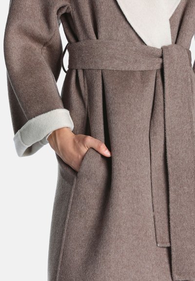 Main dans la poche d’un manteau en laine marron avec une ceinture assortie et une doublure en polaire blanche visible au col et au poignet de la manche.