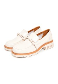 Mocassins en cuir verni blanc avec une semelle en caoutchouc épaisse, présentant une sangle décorative et des détails de couture. Texture lisse et accents beiges.