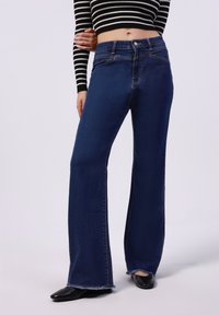 Jeans de denim de pierna ancha en azul oscuro con un dobladillo ligeramente deshilachado. Presenta un clásico diseño de cinco bolsillos y sutiles detalles de costura.