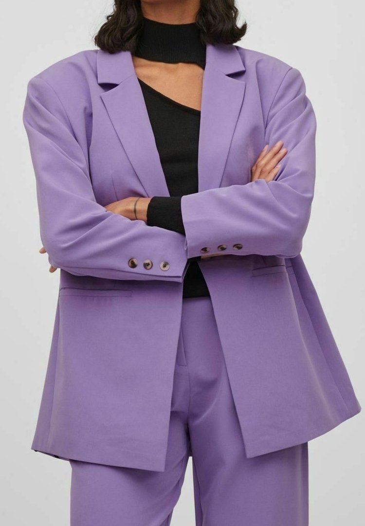 Blazer violet en tissu lisse, avec une coupe ajustée comprenant des revers, des poches avant et des poignets à boutons. Porté par-dessus un haut noir.