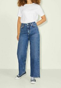 Jean en denim bleu à taille haute et jambes larges, avec un design classique à cinq poches et un ourlet effiloché, associé à un t-shirt blanc ajusté.