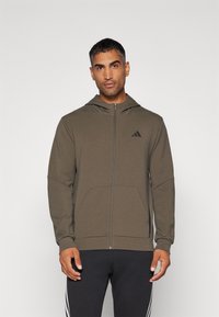 Sudadera con capucha de tela verde oliva con cierre de cremallera en el frente, bolsillos tipo canguro y un logo negro en el pecho. Textura suave, corte relajado.