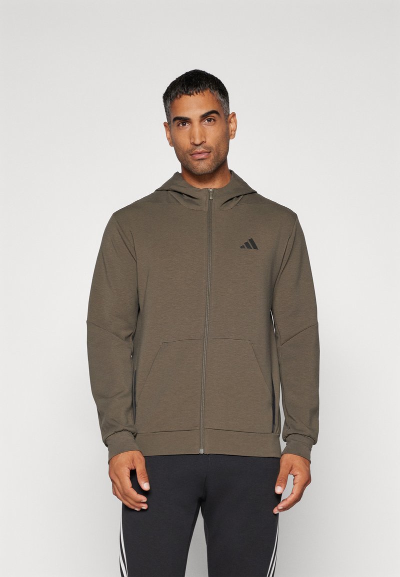 Sudadera con capucha de tela verde oliva con cierre de cremallera en el frente, bolsillos tipo canguro y un logo negro en el pecho. Textura suave, corte relajado.