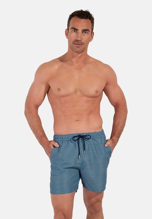 Fit man with short dark hair wearing blue patterned swim shorts with a dark drawstring, standing with hands in pockets on a white background.