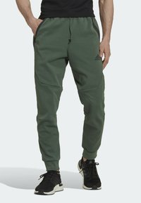 Zelené sportovní kalhoty vyrobené z měkké tkaniny, s kapsami na zip, elastickým pasem a zúženými manžetami. Doplněné černými sportovními botami.