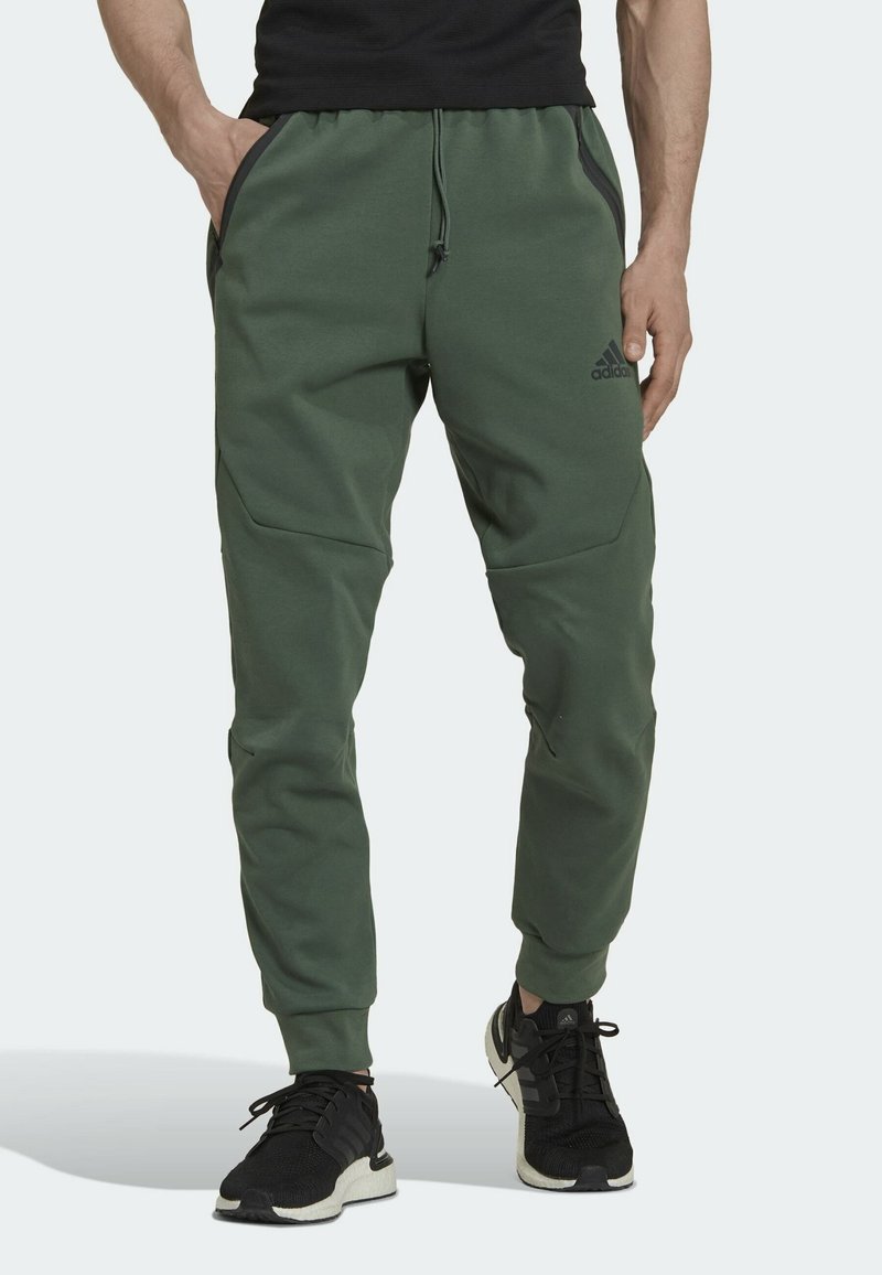 Zelené sportovní kalhoty vyrobené z měkké tkaniny, s kapsami na zip, elastickým pasem a zúženými manžetami. Doplněné černými sportovními botami.