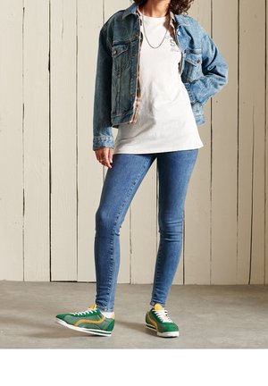 Giubbotto di denim, t-shirt bianca con grafica, jeans skinny blu, sneakers verdi con accenti gialli e suola bianca. Sfondo in pannelli di legno.