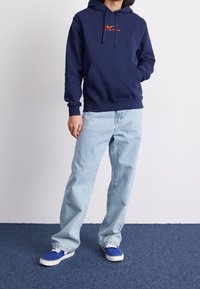 Mörkblå sweatshirt med huva och framficka, har en röd Nike-logotyp. Bärs med ljusblå lösa jeans och blå sneakers.